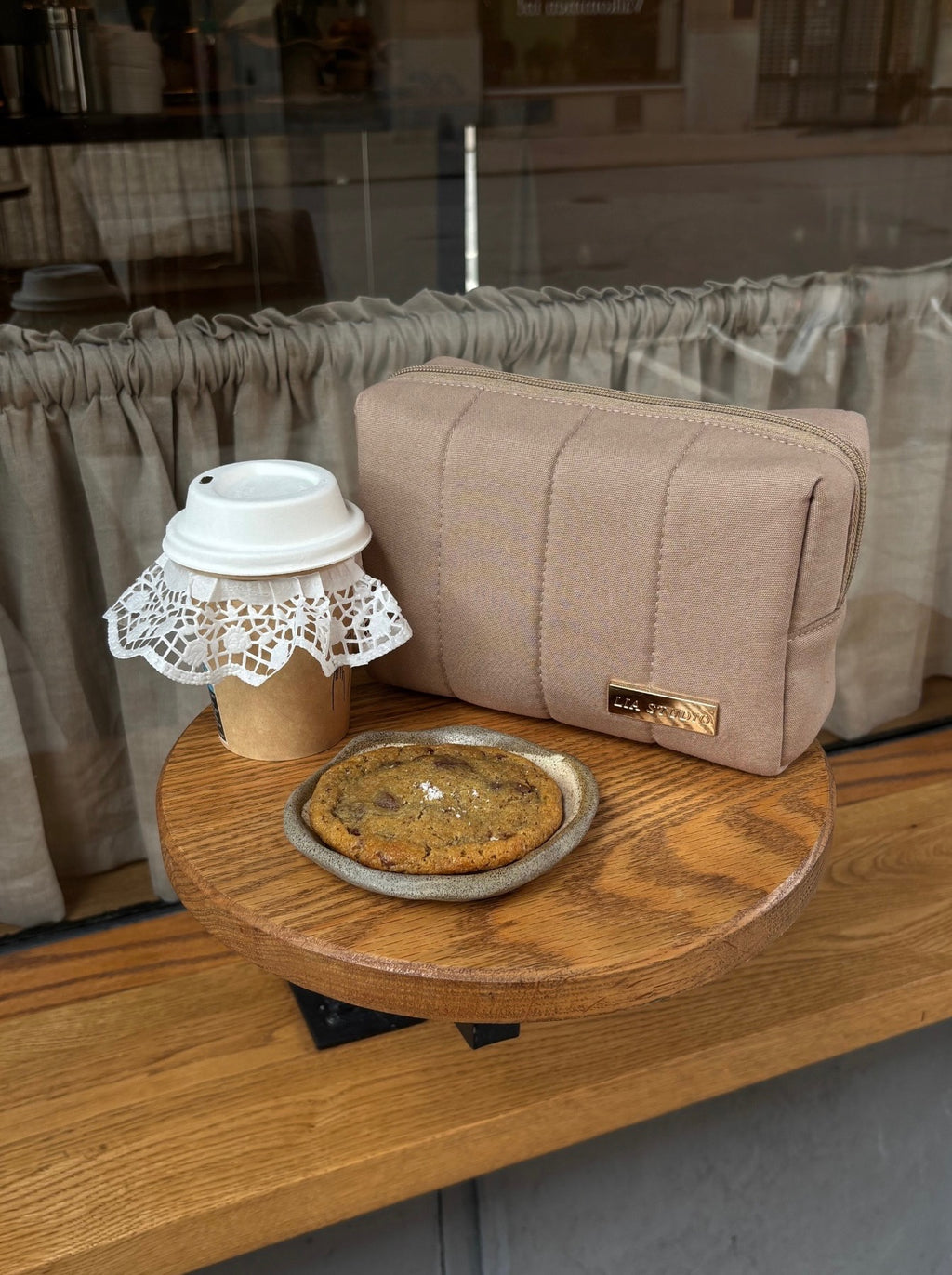 Small round wooden table with a cookie, coffee cup, and beige makeup bag in a casual setting.