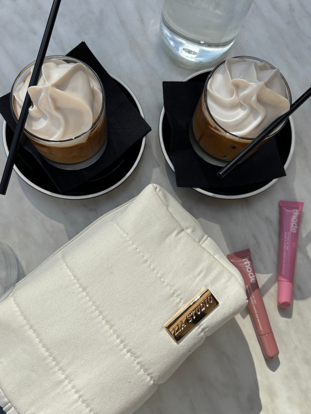 Two drinks with whipped cream, a white makeup bag with gold logo "Lia Studio" , and lip products on a light surface.