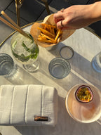 Hand reaching for fries on a glass table with drinks and white makeup bag with metalic logo "Lia studio"