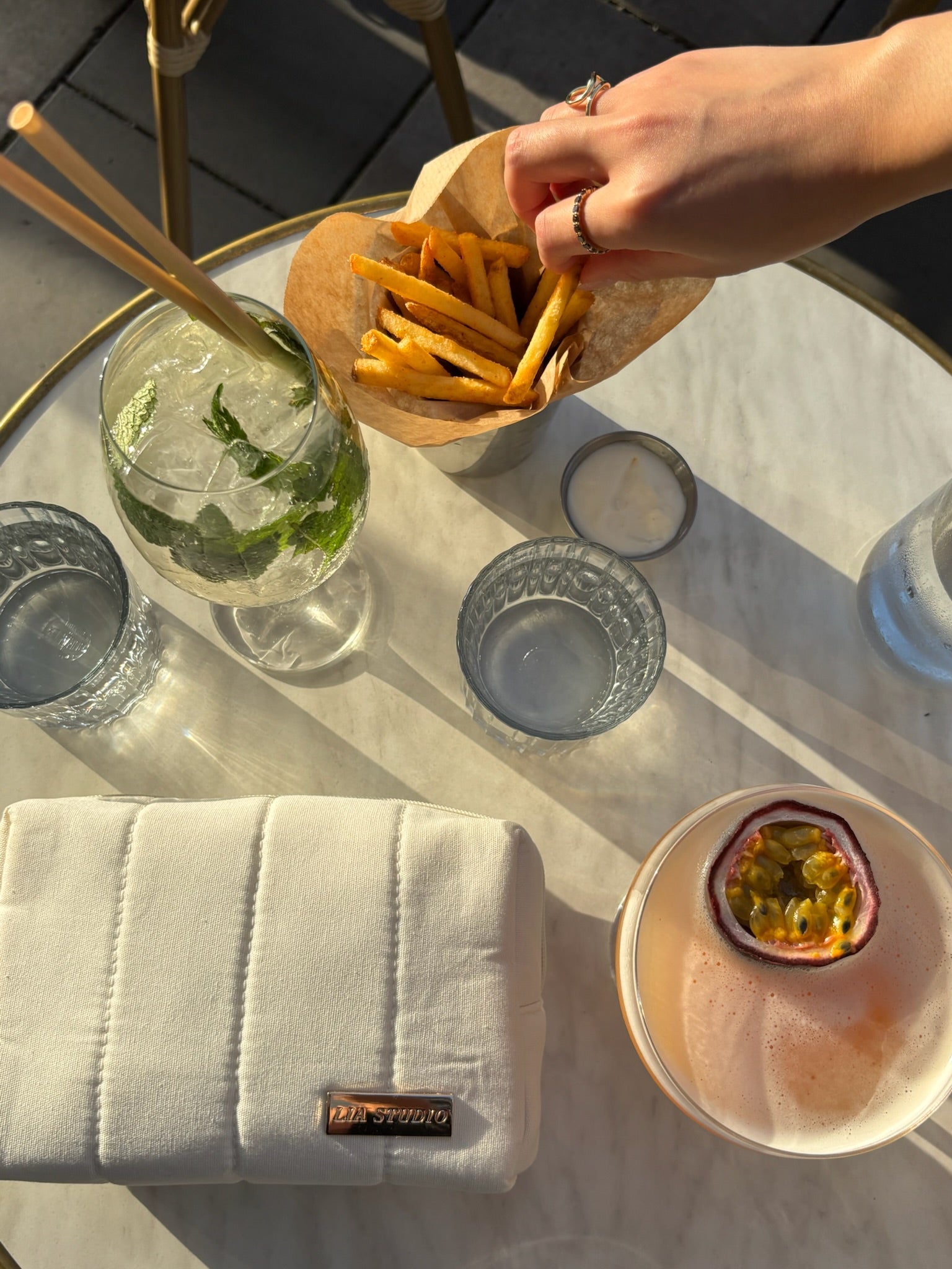 Hand reaching for fries on a glass table with drinks and white makeup bag with metalic logo "Lia studio"