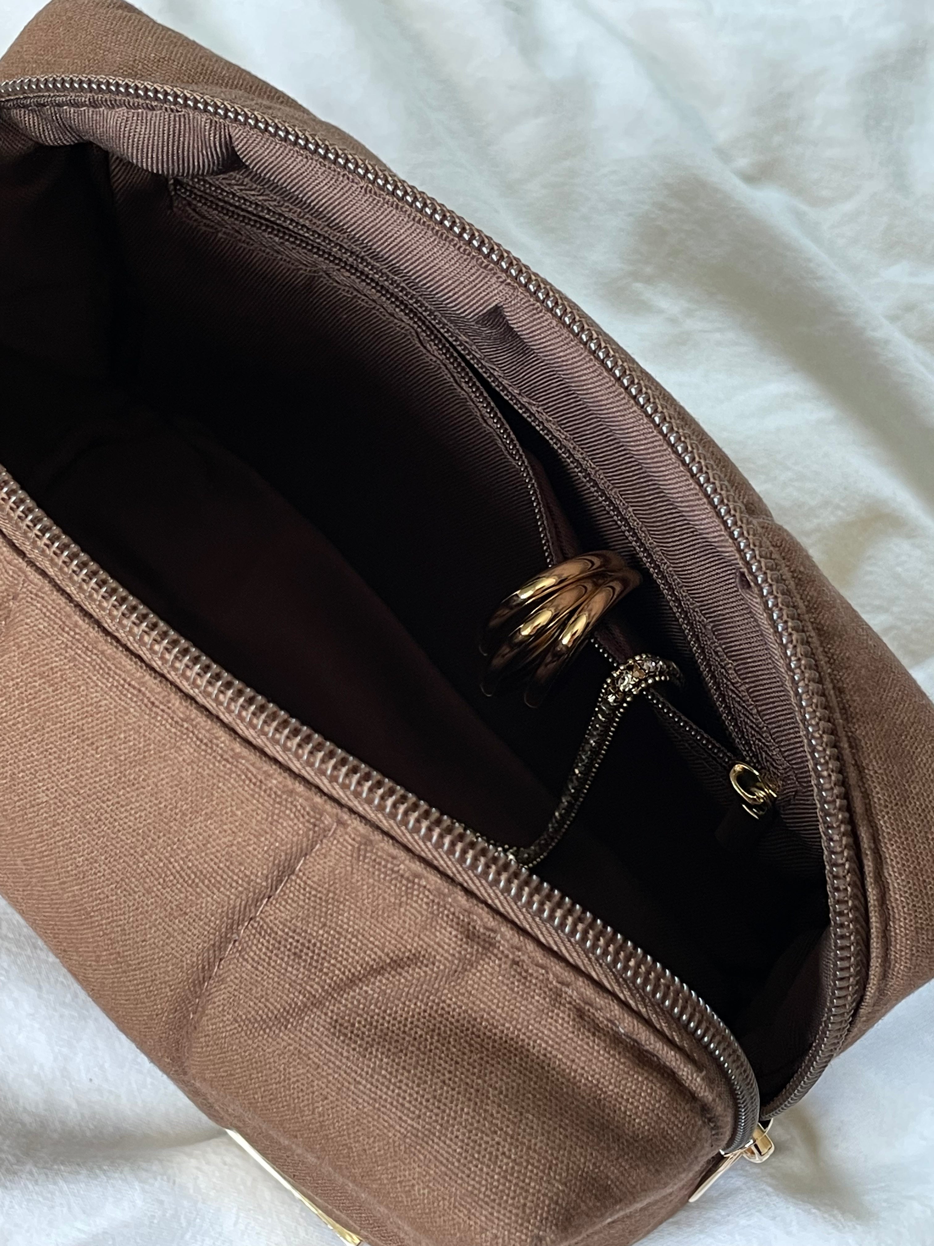 Brown makeup bag with a visible inner pocket with gold jewelry inside on a white background