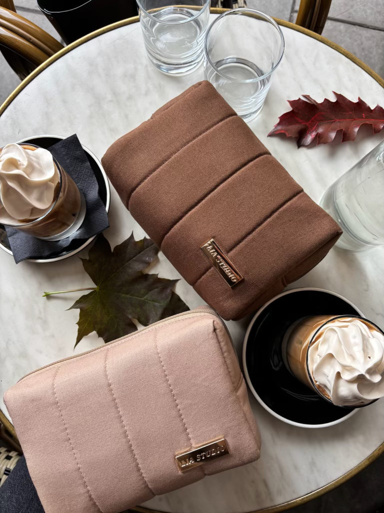Two makeup bags, one brown and one beige, on a table with coffee and water.