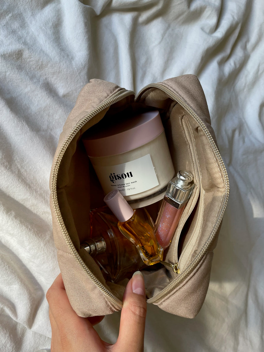 hand holding beige makeup bag in the color chai open on bed with products inside