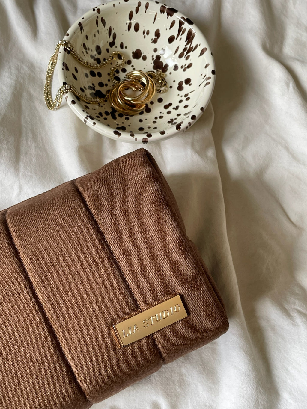 brown makeup bag tonka on white textured fabric by bowl with gold jewelry
